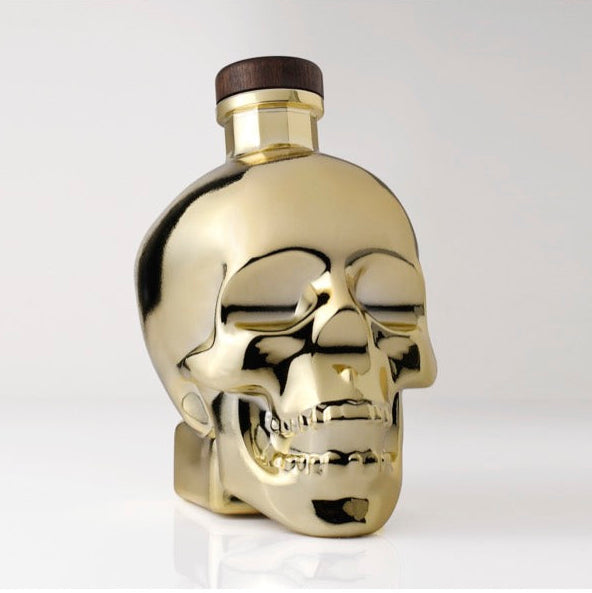 A metallic gold skull-shaped bottle with a dark brown cap, shown on a light gray background.