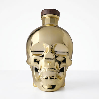 A metallic gold skull-shaped bottle with a dark brown wooden cap, shown against a plain white background.